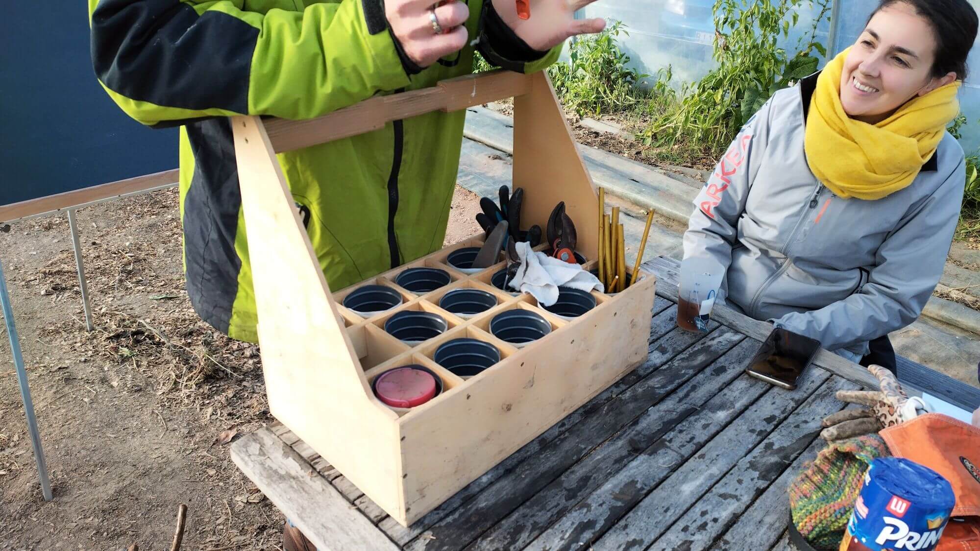 Plan rapproché sur le kit à bouture de Benjamin : une structure en bois léger composé de cases, pour caler les sécateurs, le petit matériel, et les boutures (dans une moitiée de bouteille en plastique avec de l'eau au fond)